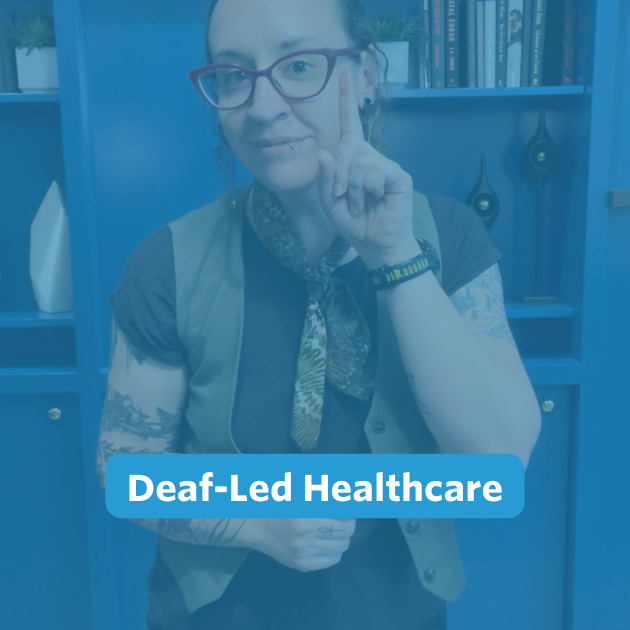 A nonbinary person with long, curly brown hair pulled back stands in front of the camera. Behind them is a blue built-in bookshelf wall. They are wearing black pants, a black shirt layered with a forest green vest, a mixed green neck bandana, and a pair of gold and green earrings. They are signing, 'deaf'.